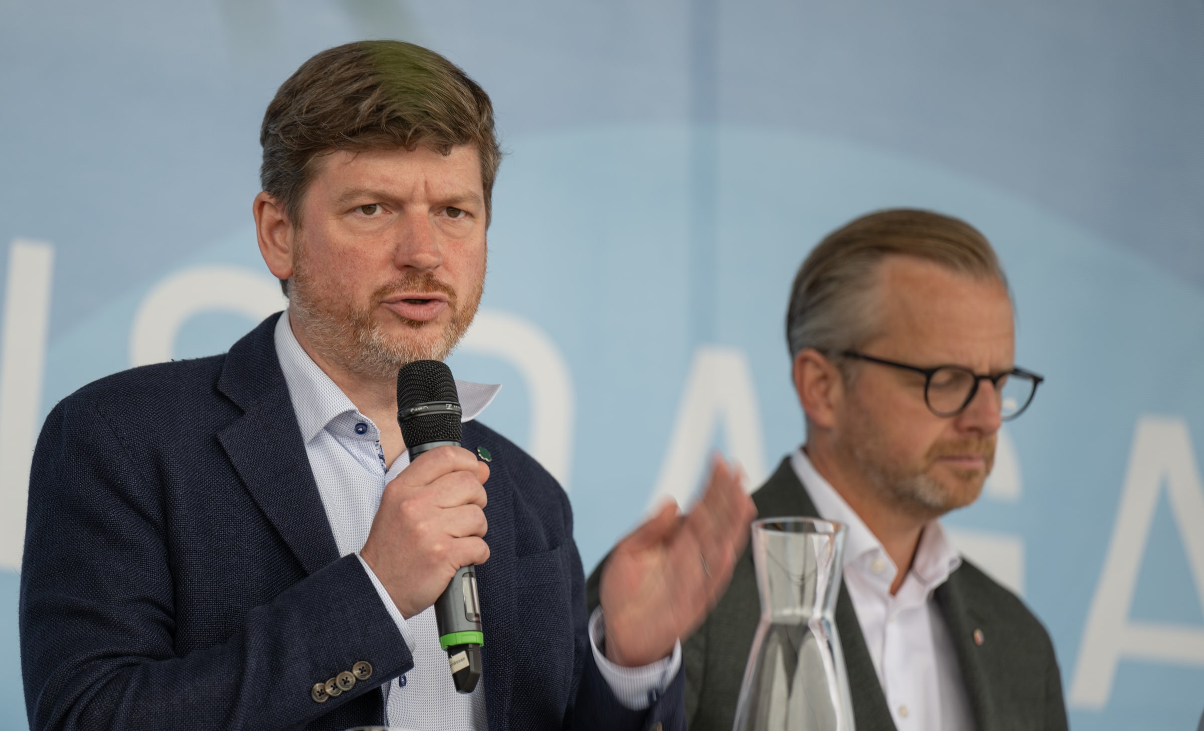 Martin Ådahl speaking into a handheld microphone and gesturing on a public debate stage, with another participant behind him.