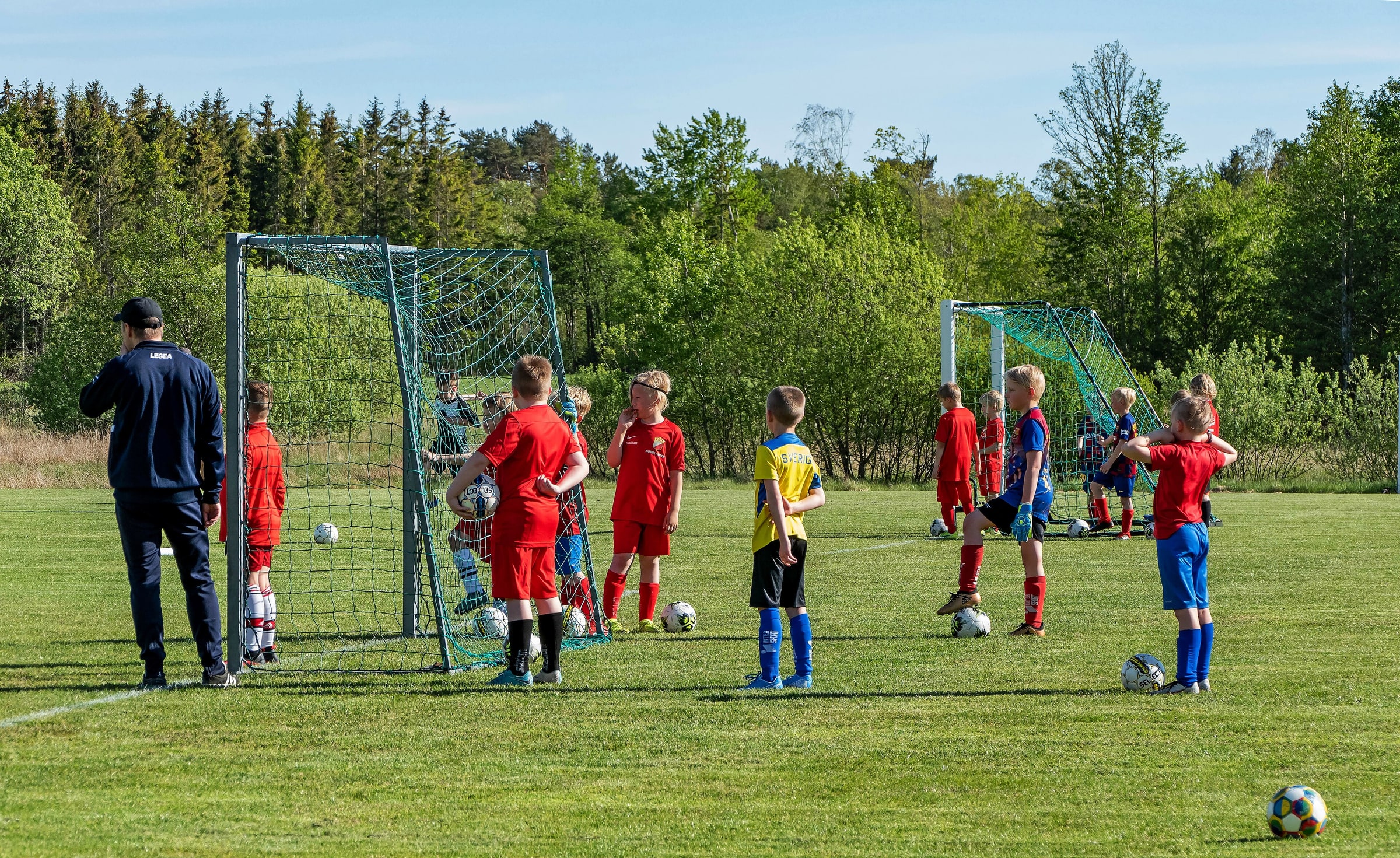 Children and a coach gather with footballs around two small goals on a Swedish grass pitch.