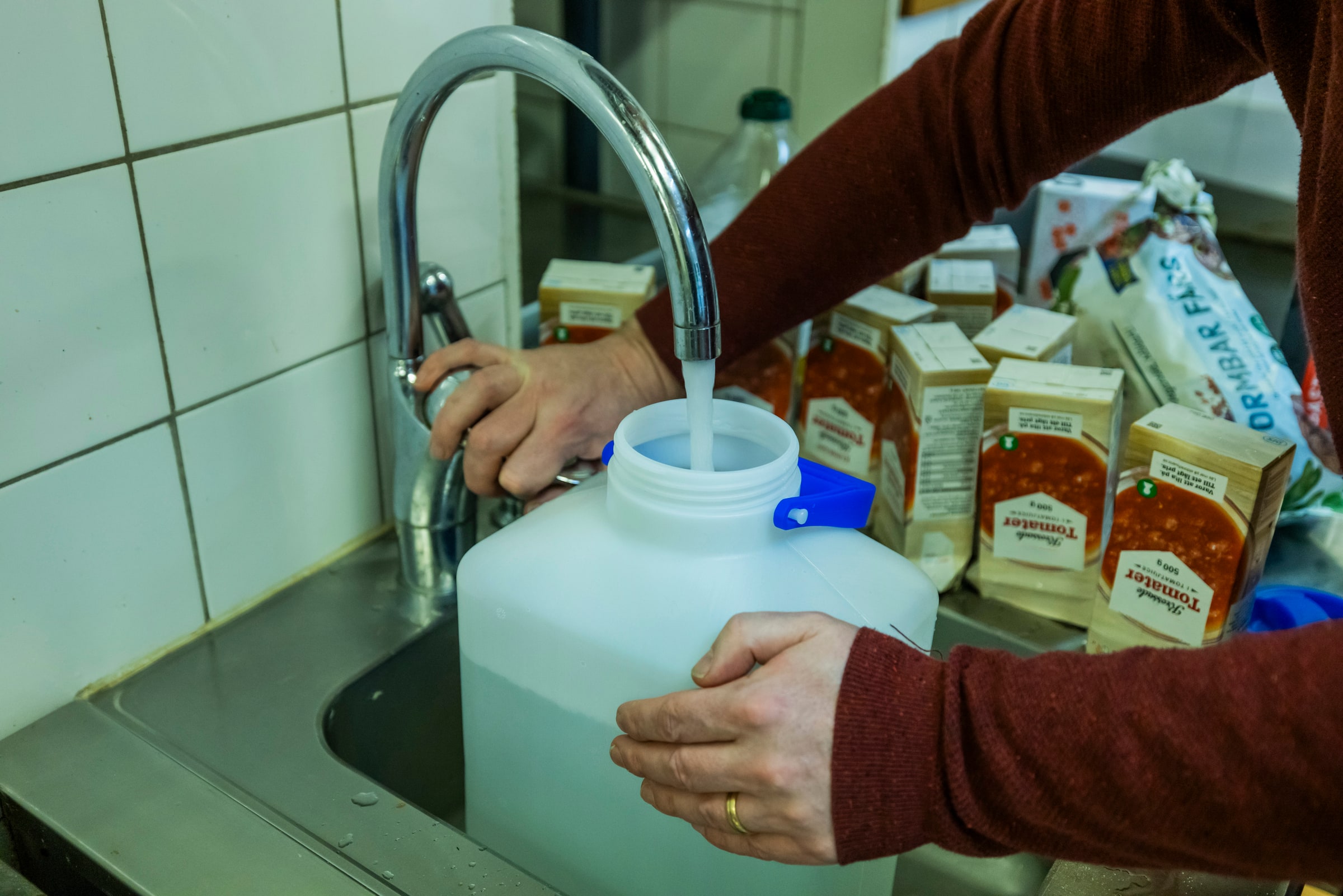 A person fills a white water container at a kitchen tap beside stored food.