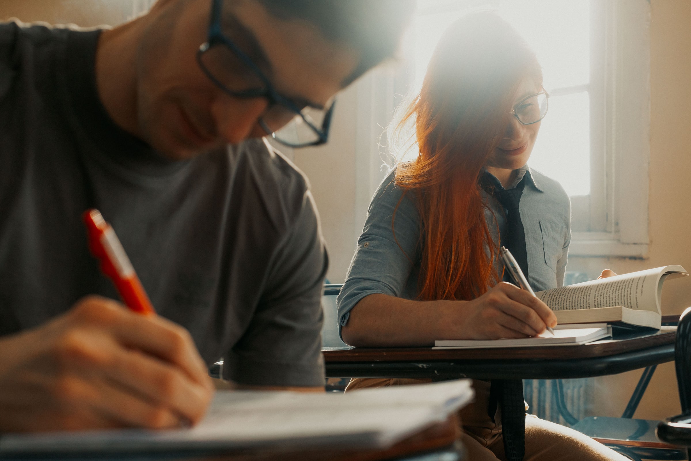 Two adult students write notes at classroom desks, with an open book beside them.