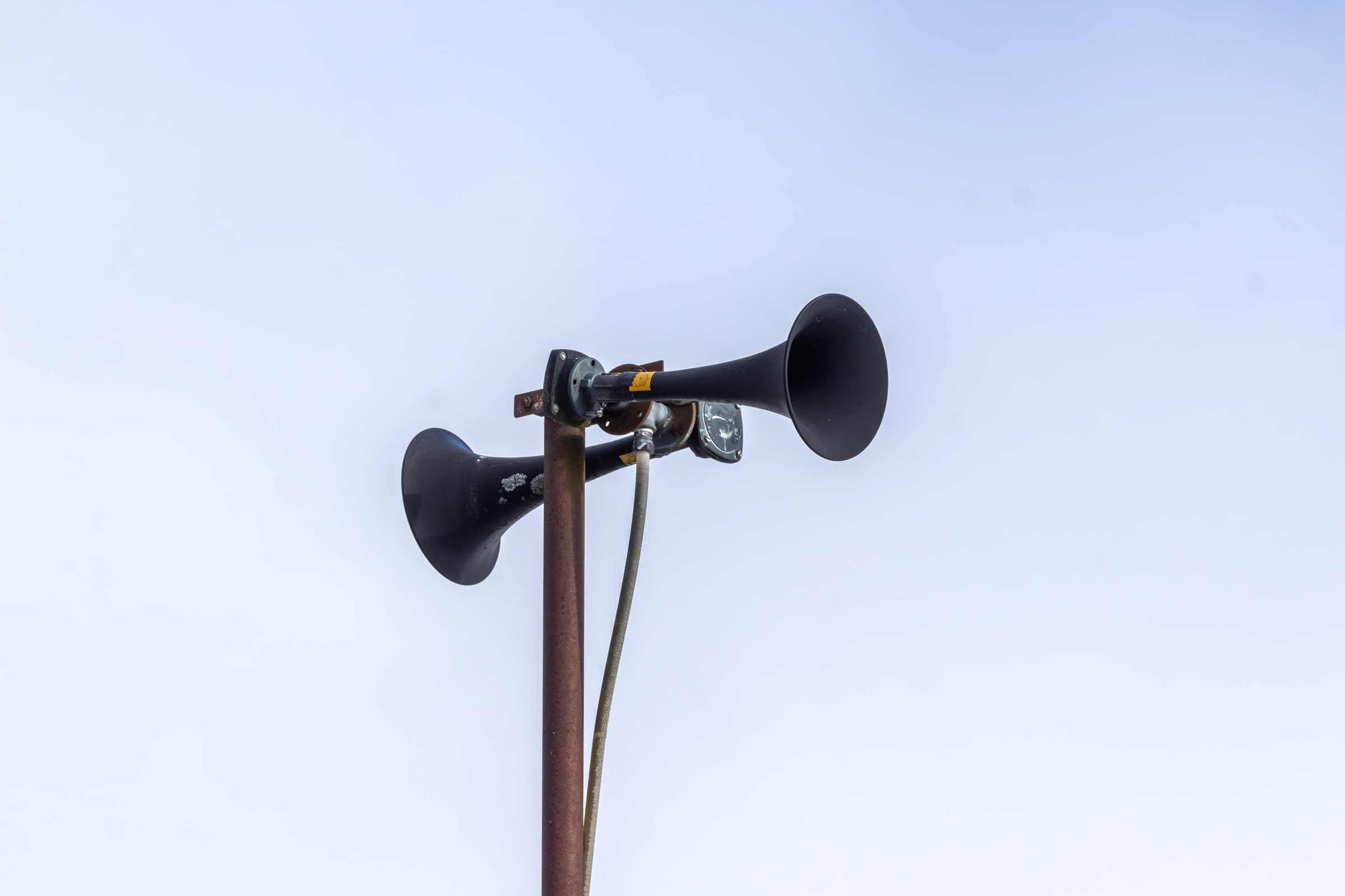 Two black outdoor public-warning horns mounted on a metal pole against trees and sky.
