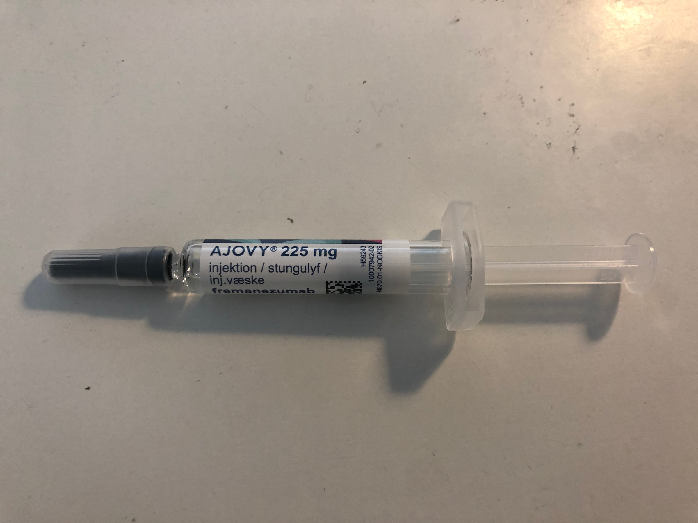 A complete prefilled syringe labelled AJOVY 225 mg rests on a neutral surface.