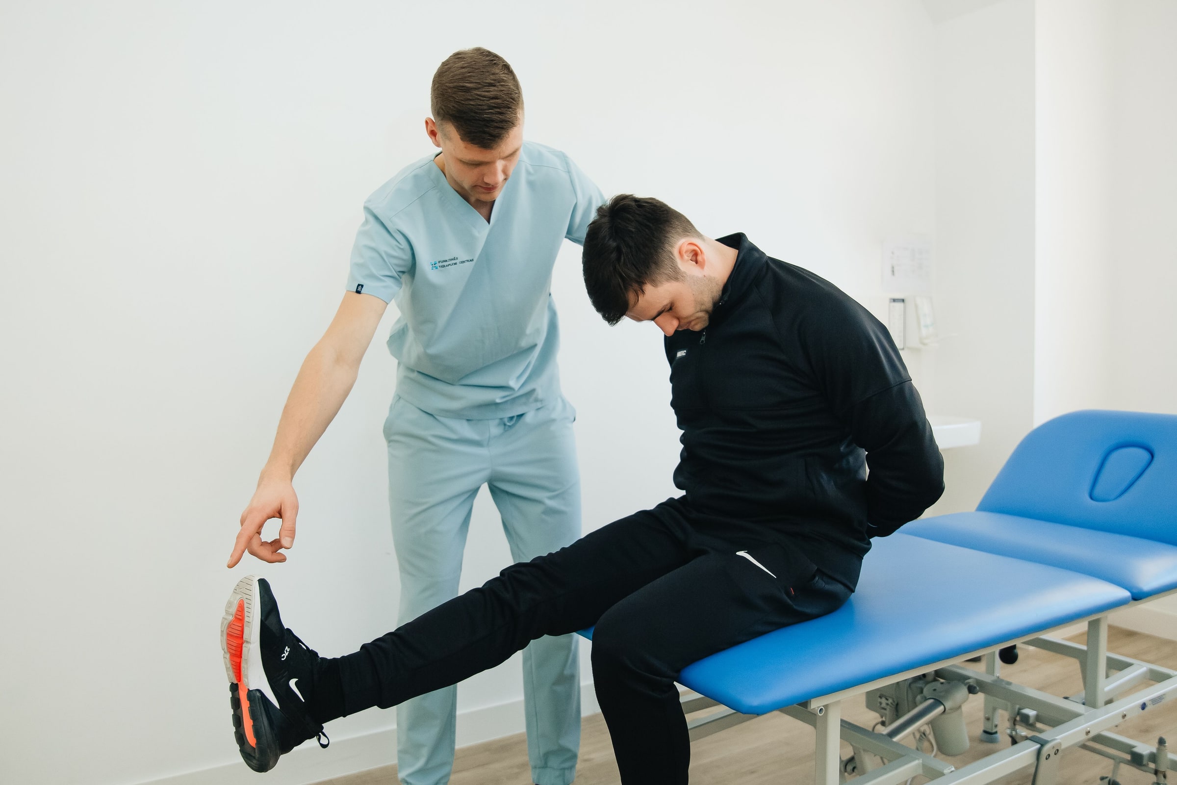 A physiotherapist guides a seated patient extending one leg in a clinical rehabilitation room.