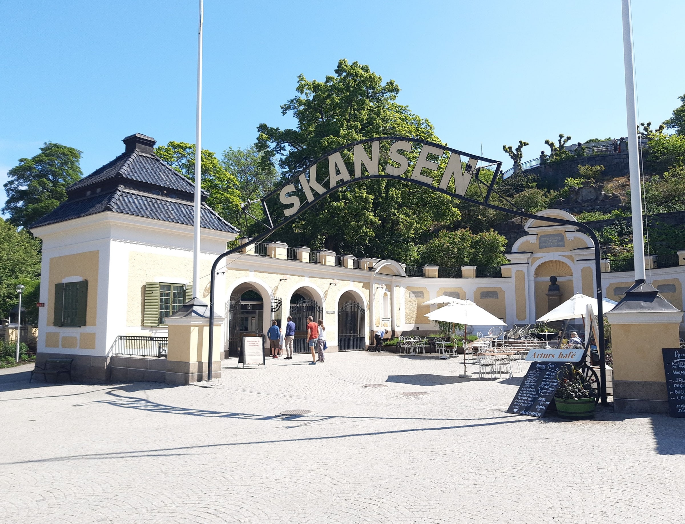 Skansen’s cream and yellow entrance building, arched sign and paved forecourt, with distant visitors at the side.