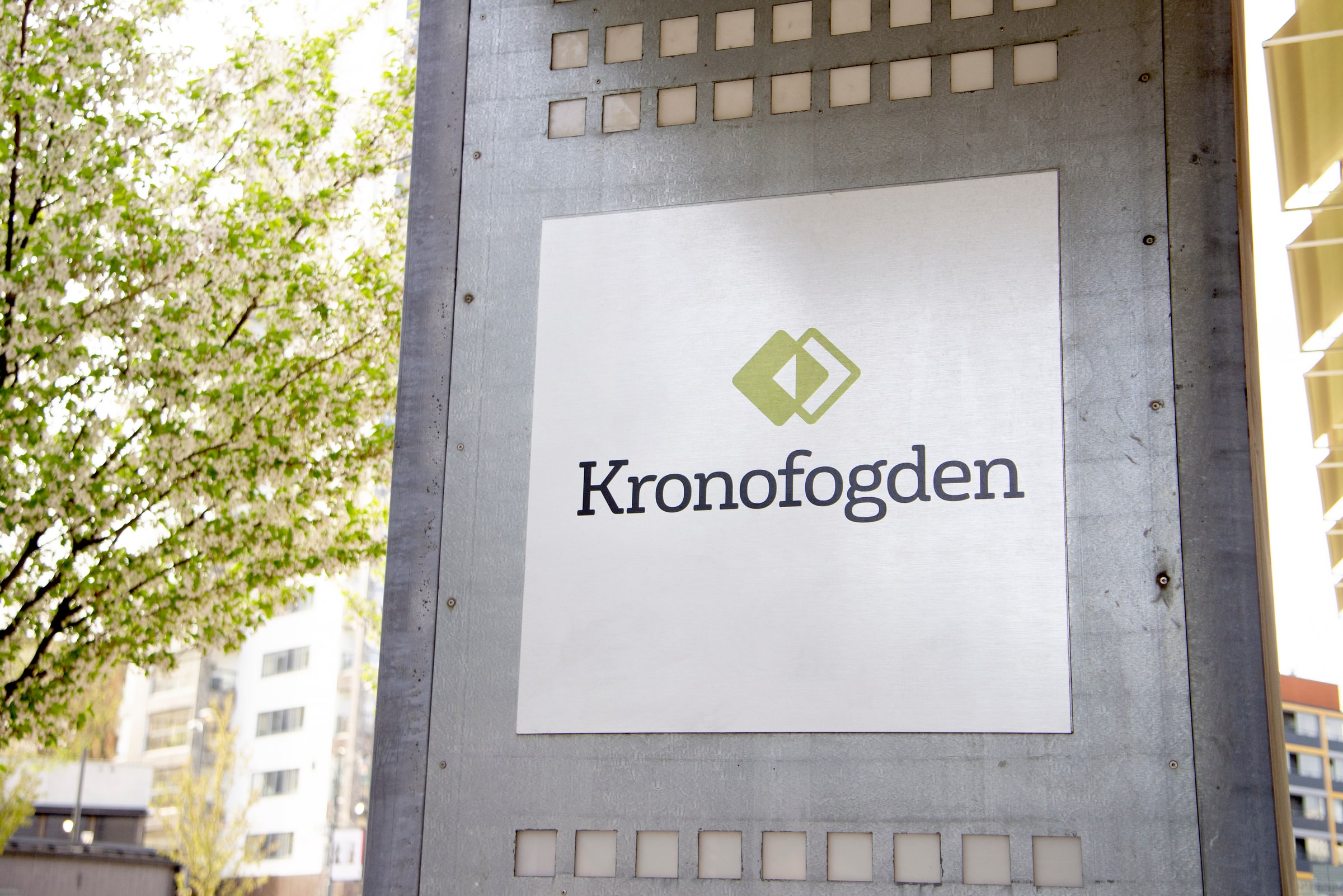 A Kronofogden sign mounted on an exterior wall beside a flowering tree in daylight.