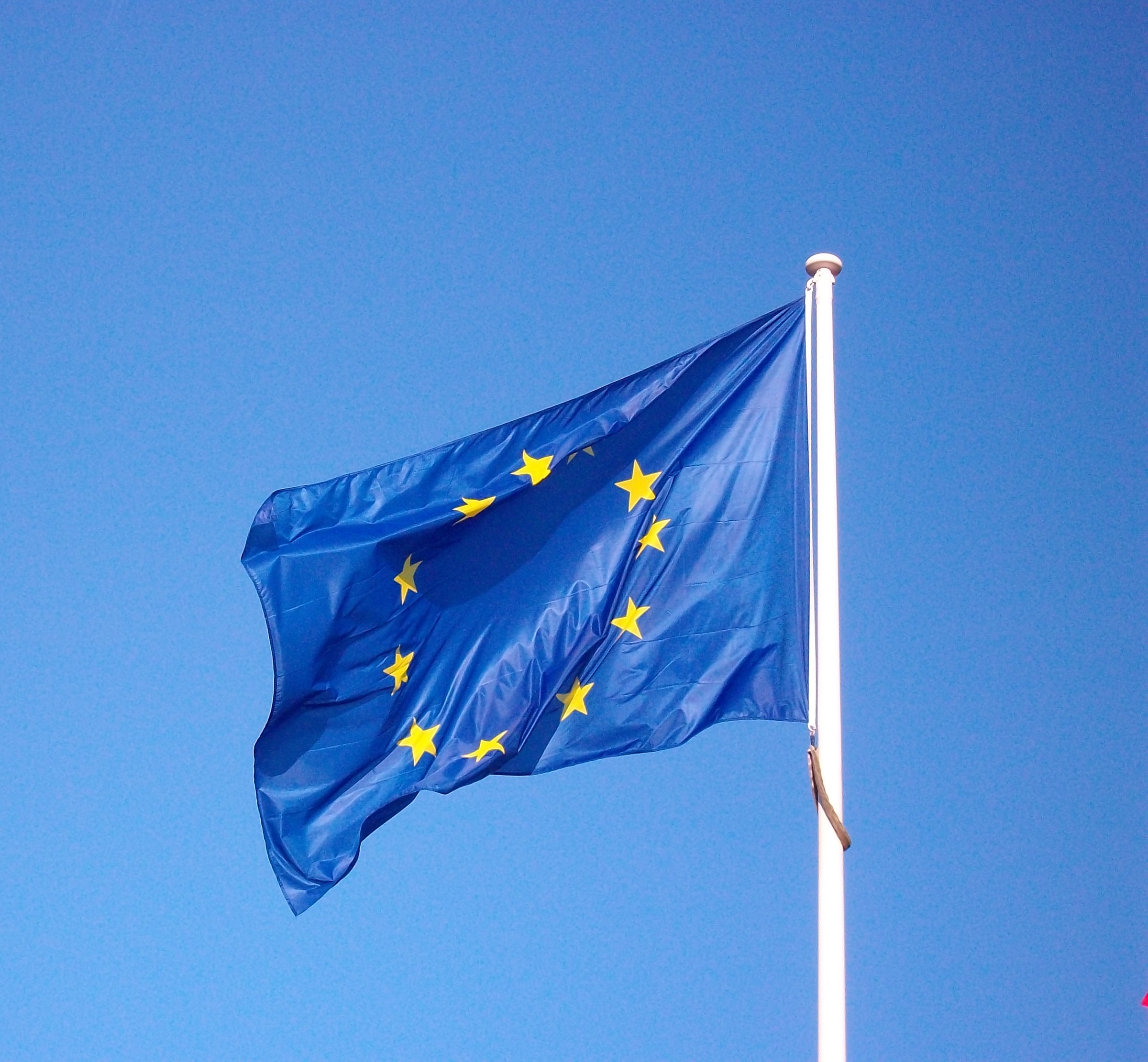 A blue European Union flag with gold stars flying against a blue sky.