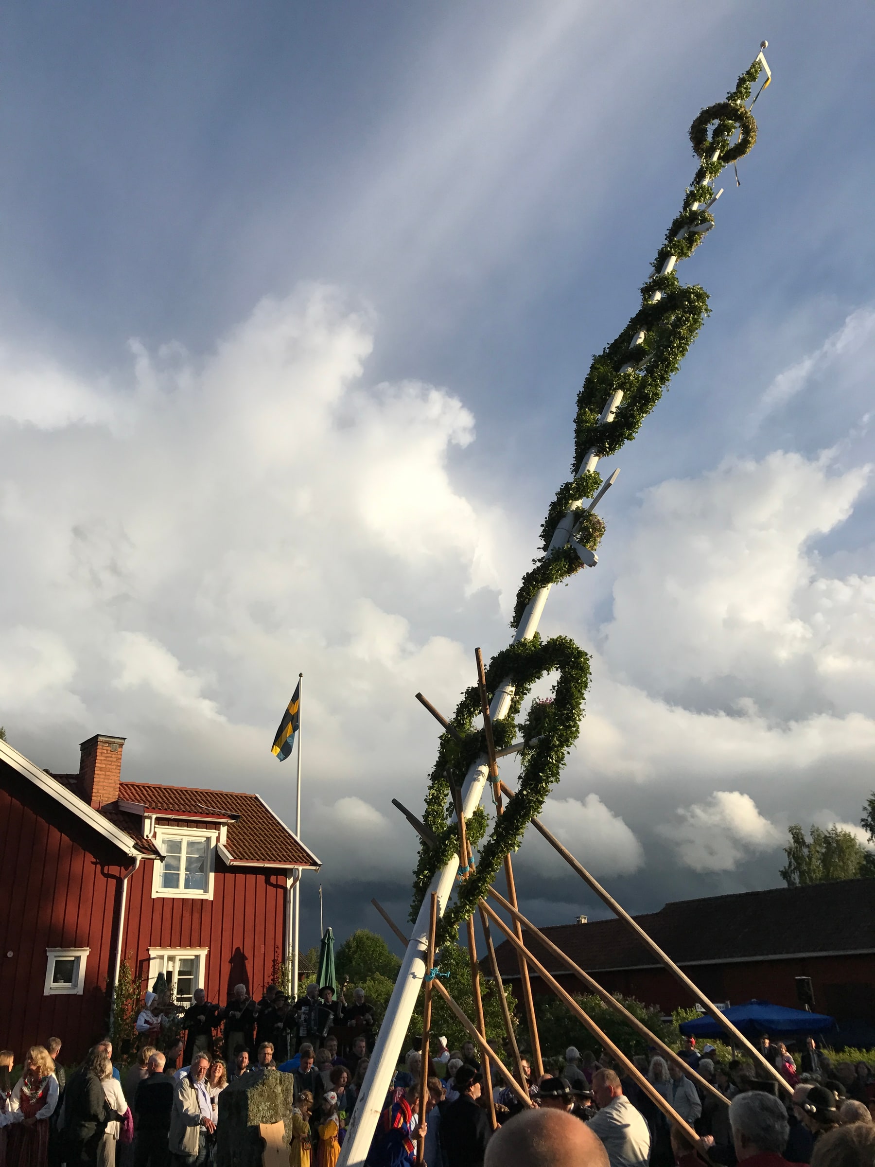 People gather around a decorated Midsummer maypole beside red wooden buildings in Rälta, Sweden.