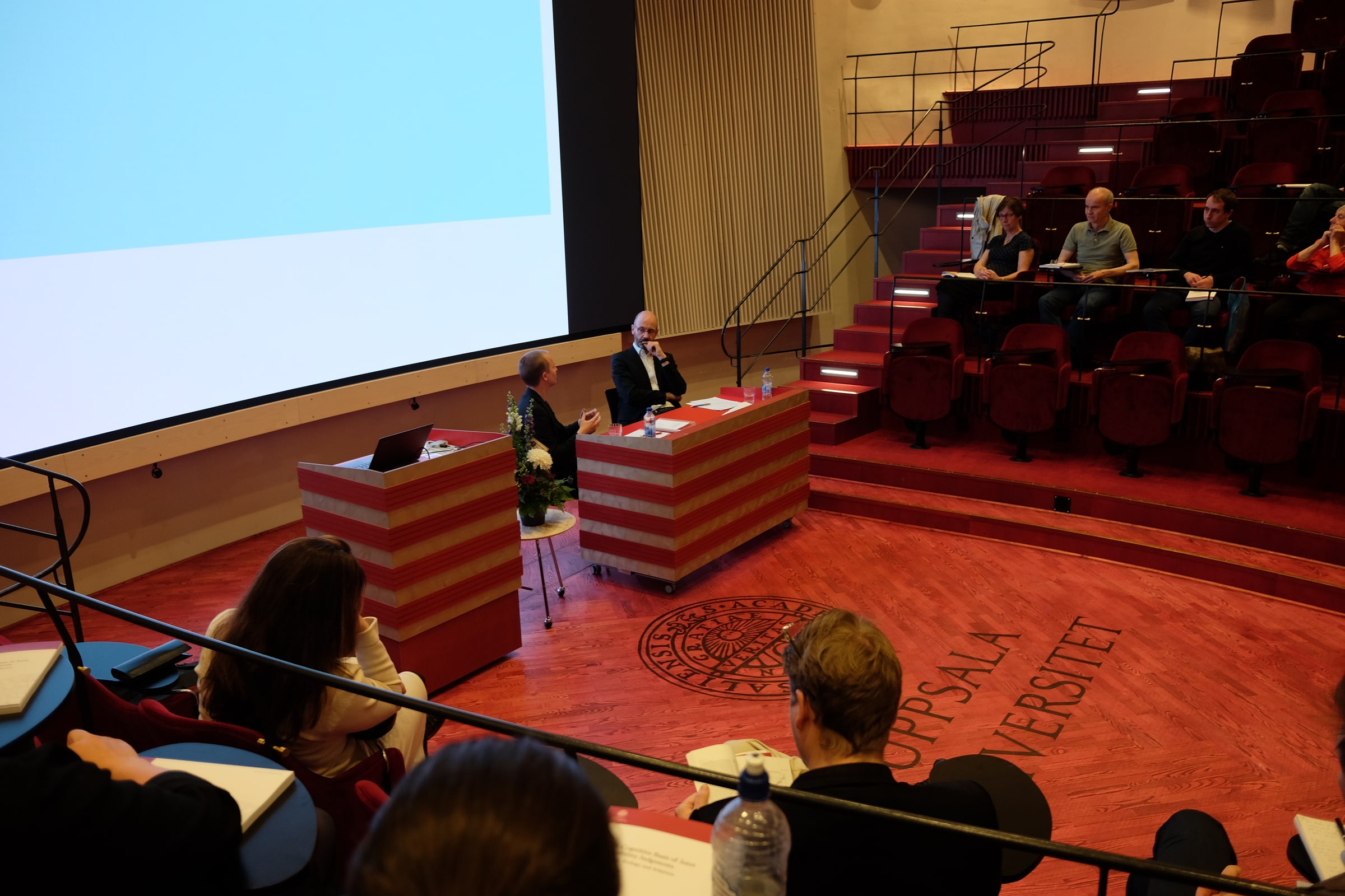 A doctoral respondent gestures towards the examiner across two lecterns, with an audience seated around them at Uppsala University.