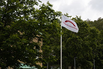 A white Migrationsverket flag flying outside against green trees.